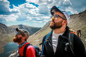 Two men standing side by side mountainside, staring up into the sunny sky wearing NOZ reef safe and cruelty-free SPF in two different neon colors. 